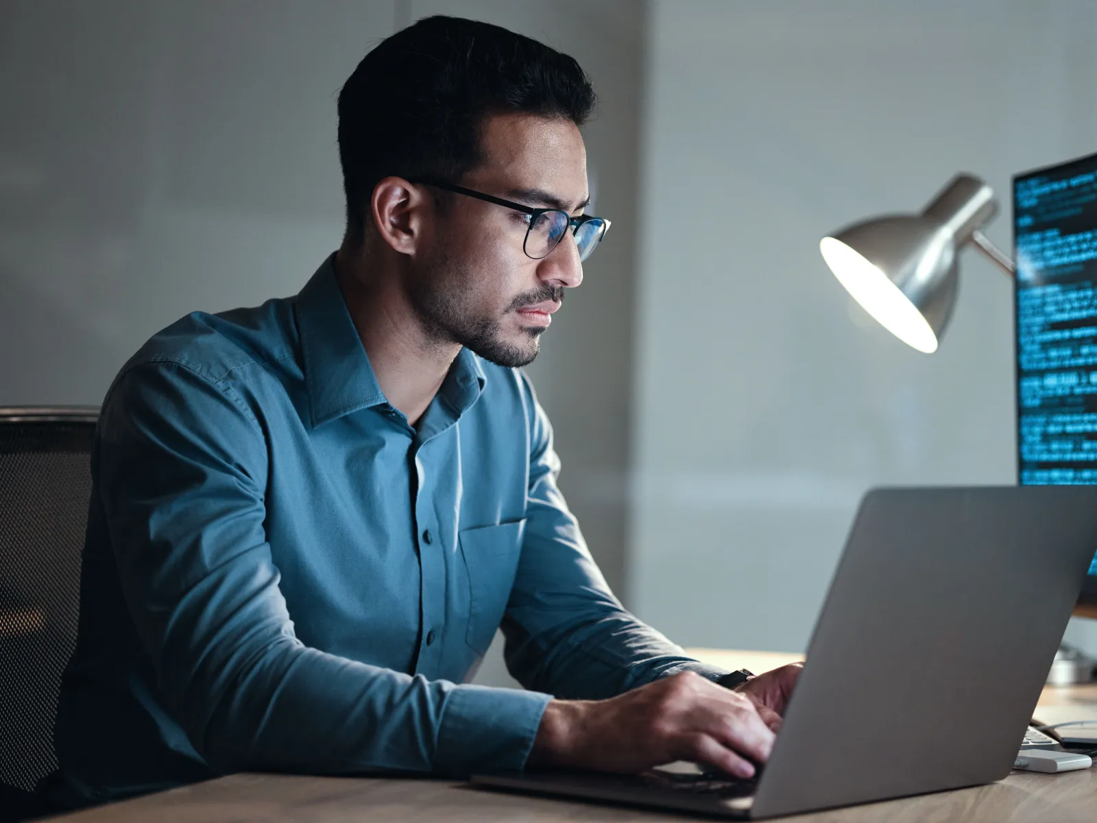 A focused man in glasses typing on a laptop in a modern workspace with a glowing monitor.