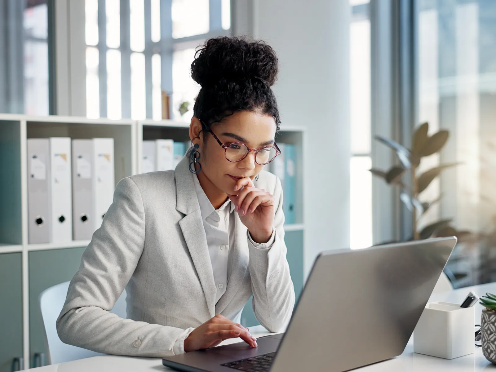 Professional woman in gray blazer working on laptop in modern office setting with natural light.