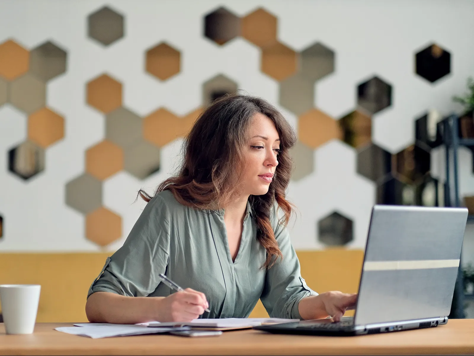A woman working on a laptop, taking notes at a desk with a geometric wall design.