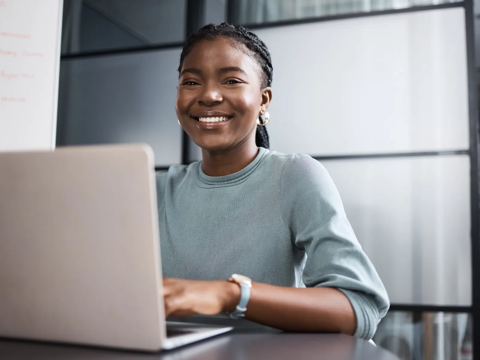 Smiling woman working on a laptop in a modern office setting, with natural light and glass backdrop.