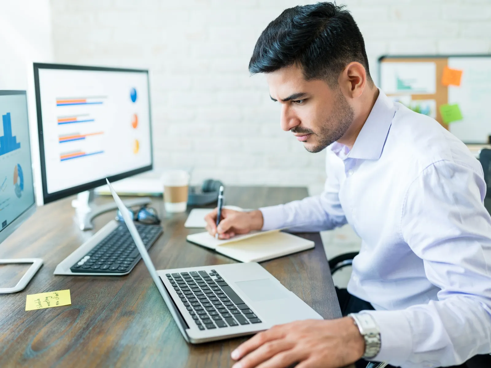 A businessman working at a desk with two monitors, taking notes while using a laptop.