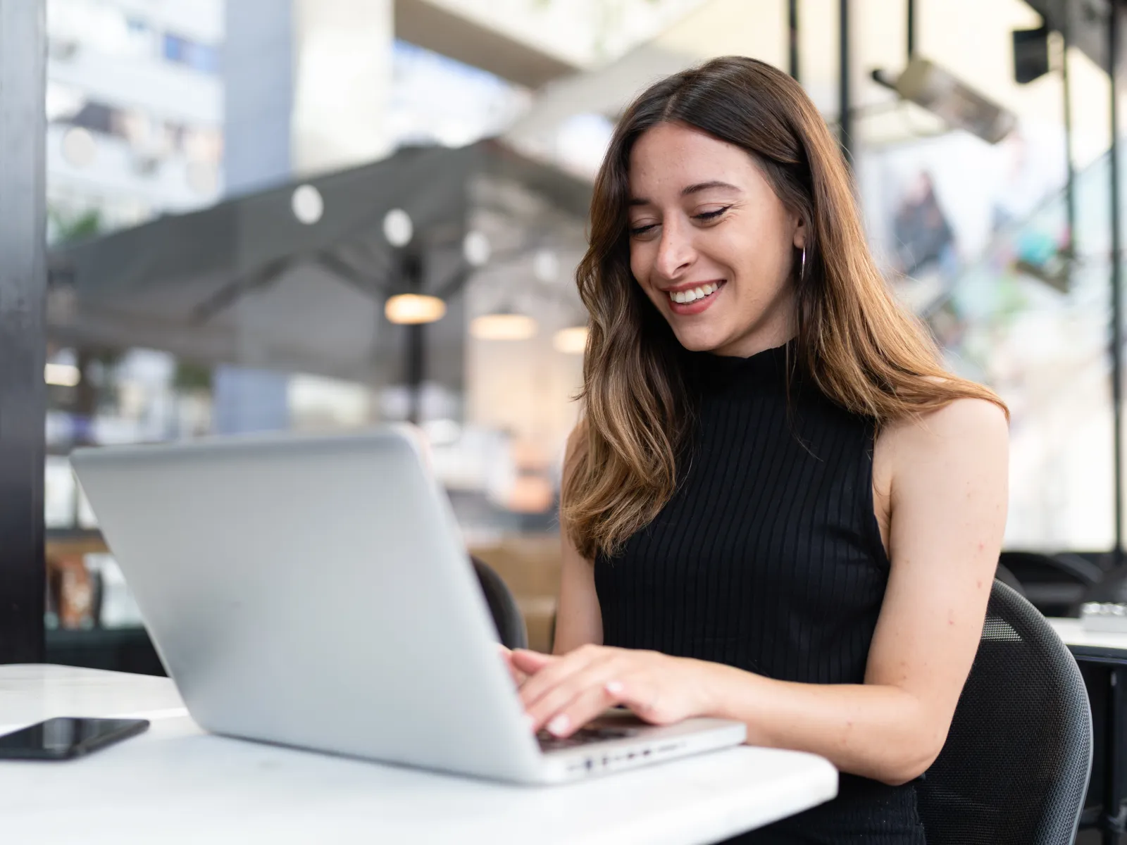 Smiling woman working on laptop at a cafe table, modern environment with soft natural light.