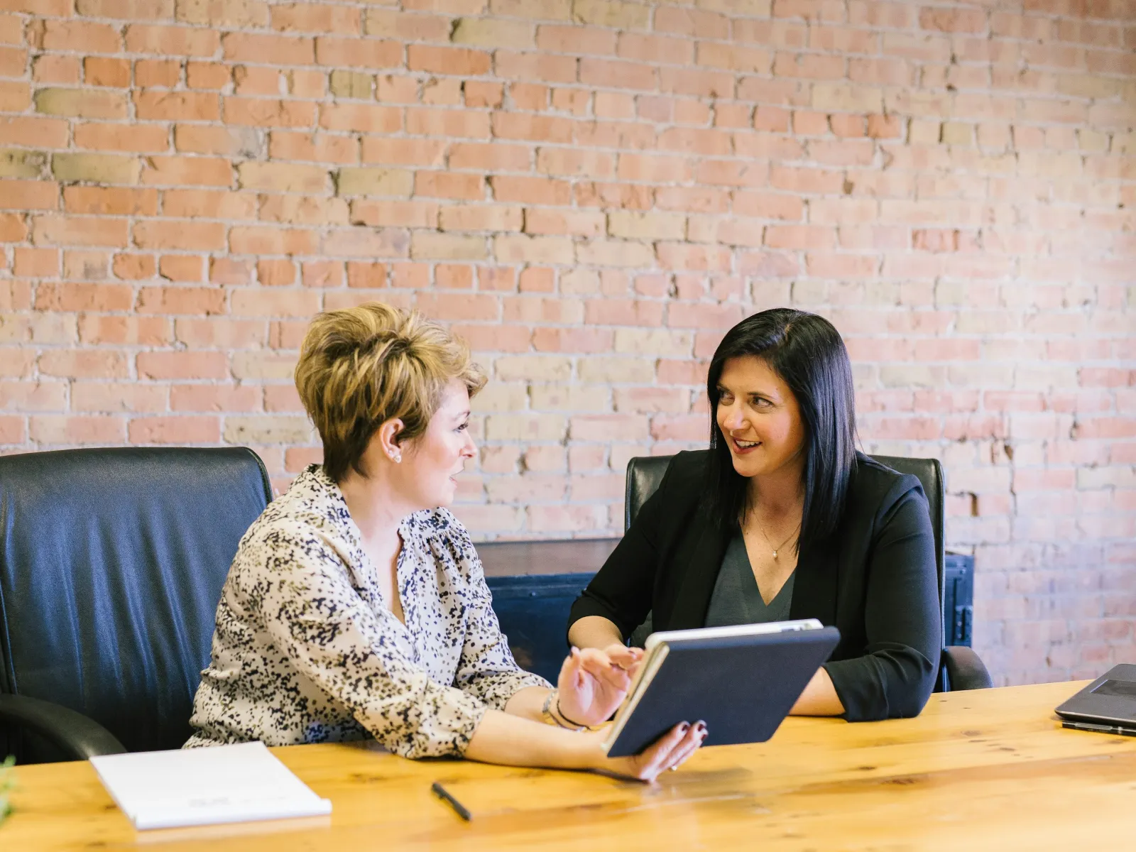 Two businesswomen discussing over a tablet in a modern office setting with a brick wall backdrop.