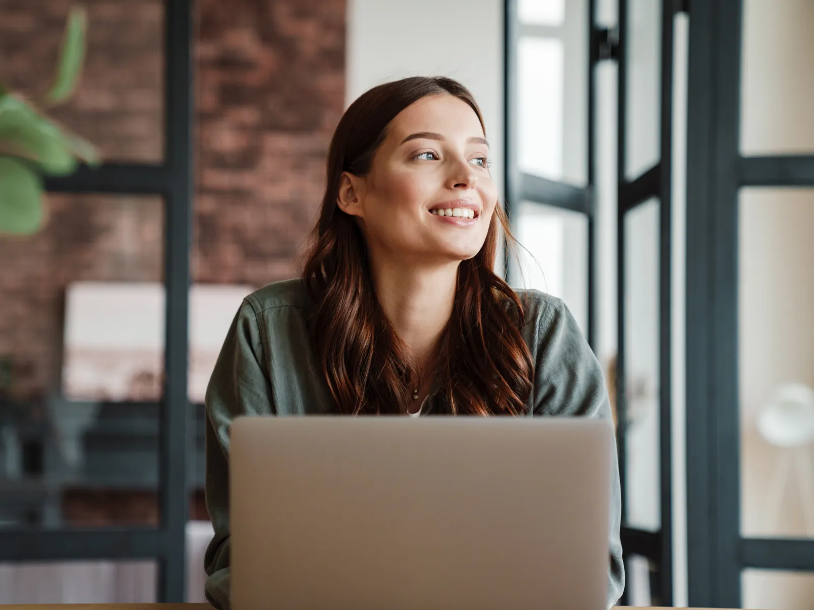 Smiling woman sitting at a desk with a laptop, surrounded by modern interior decor.