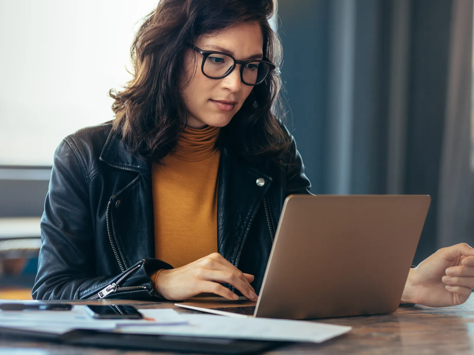 Woman in a leather jacket working on a laptop at a desk with documents and a notepad.