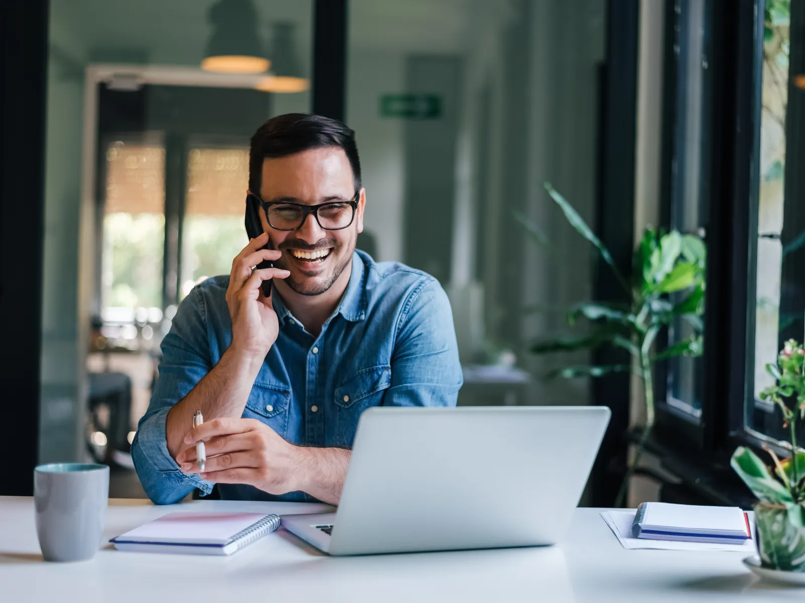 Smiling man on phone at desk with laptop, notebook, coffee, and plants in a bright workspace.