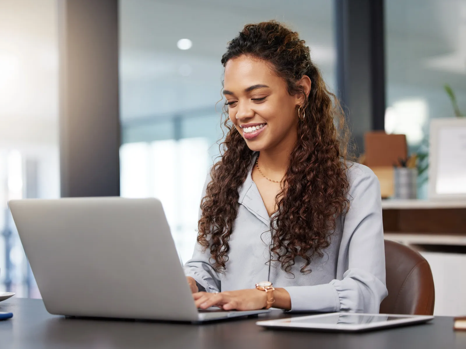 Smiling woman working on a laptop in a modern office setting, with a coffee cup nearby.