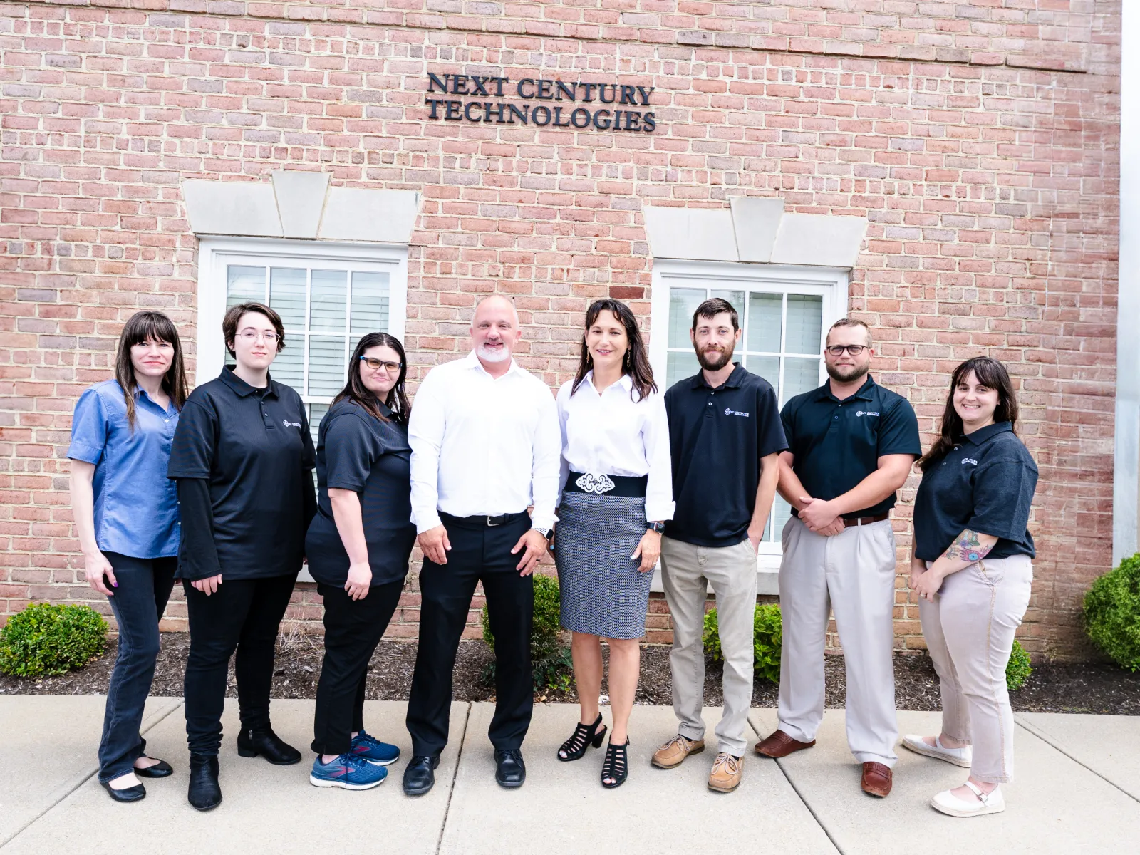 Eight professionals standing in front of Next Century Technologies office building with brick facade and windows.