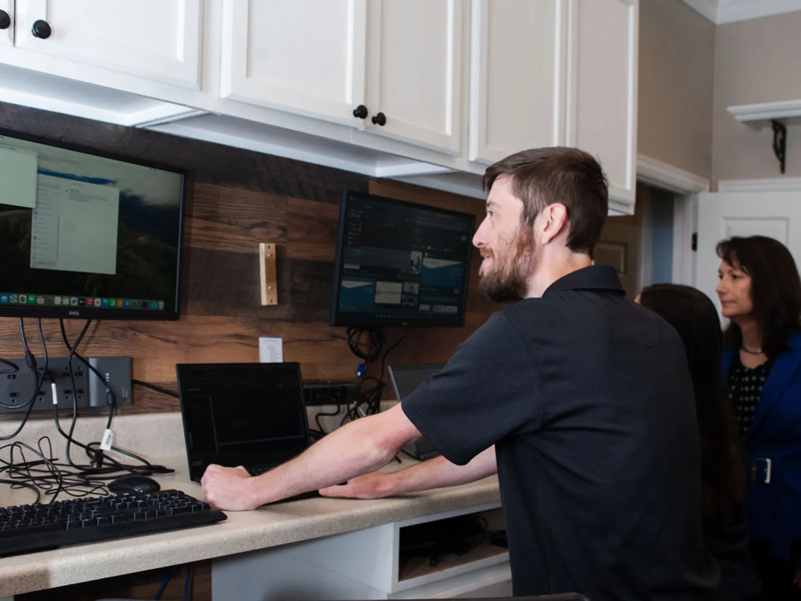 Man interacting with dual monitors and a keyboard in a modern workspace while two women observe.
