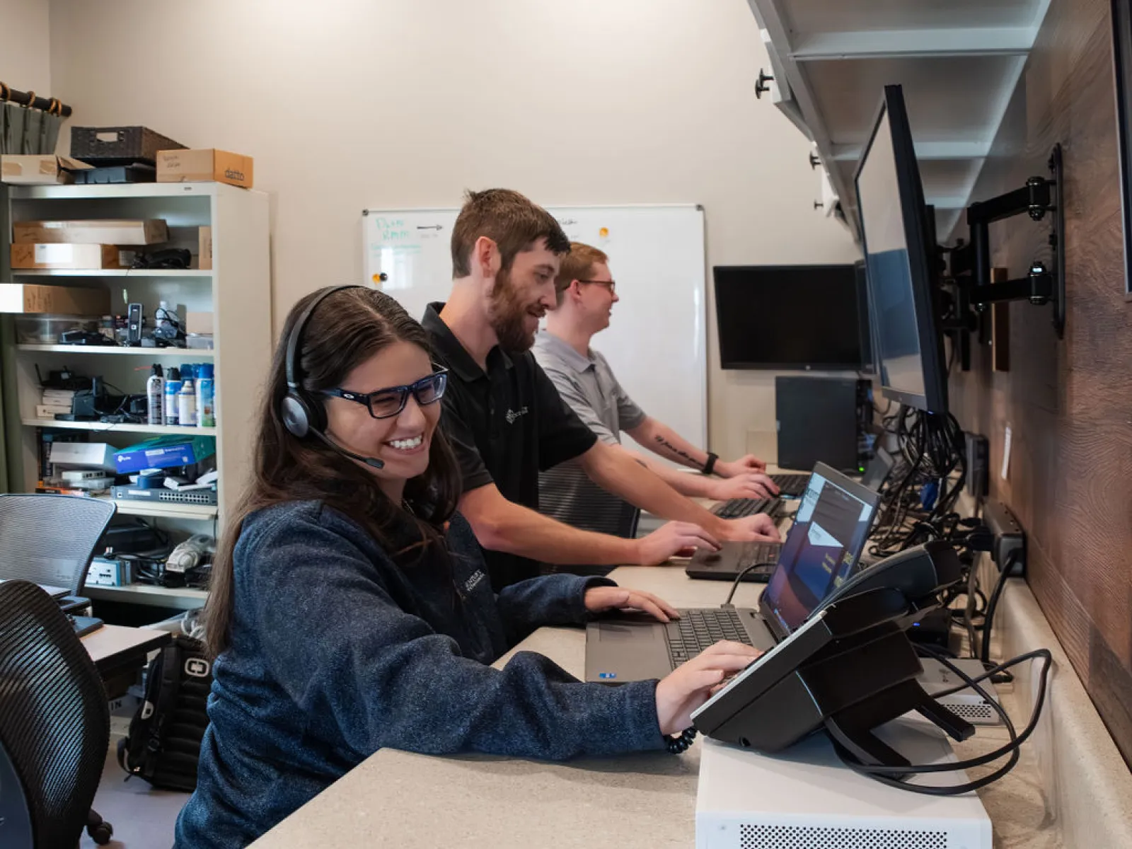 Three smiling customer service agents working at computer stations with headsets in a modern office environment.
