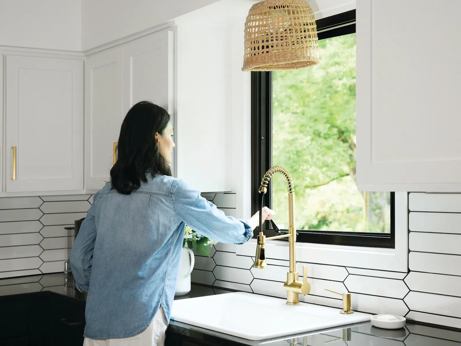 Woman in denim shirt opening a window above a modern kitchen sink with gold faucet and hexagonal tile backsplash.