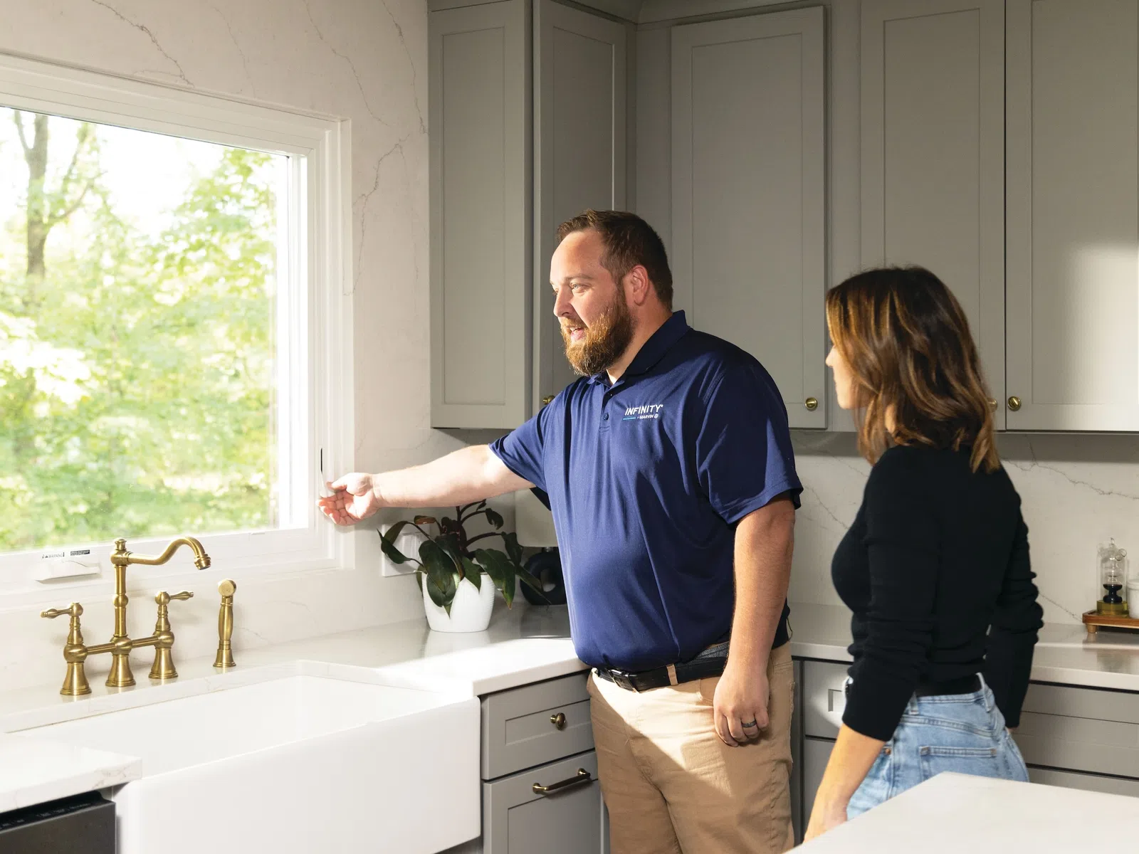 Technician in blue shirt shows woman how to open kitchen window above white farmhouse sink with brass faucet.
