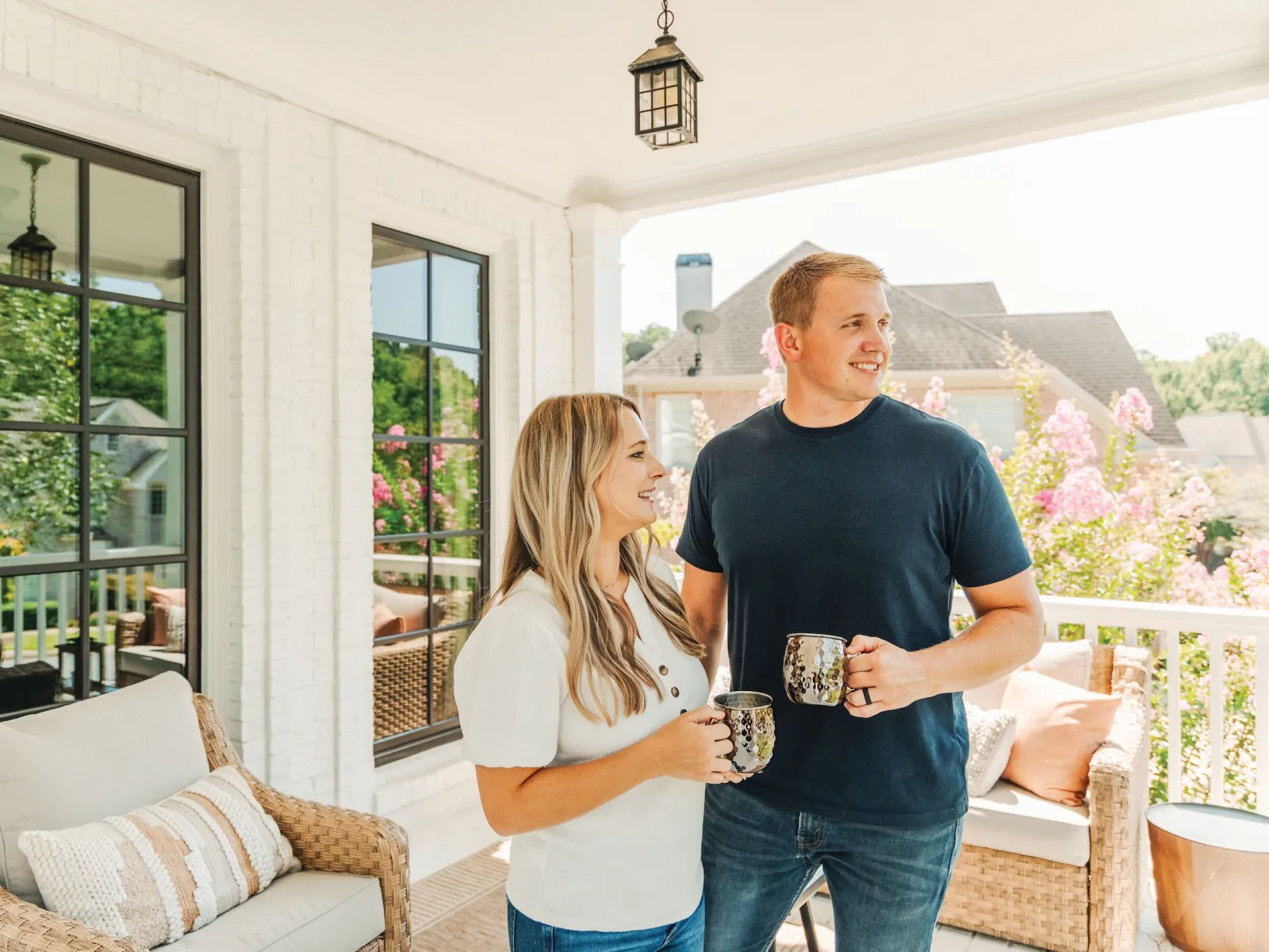 Couple enjoying coffee on a bright porch with white brick walls and wicker furniture on a sunny day.