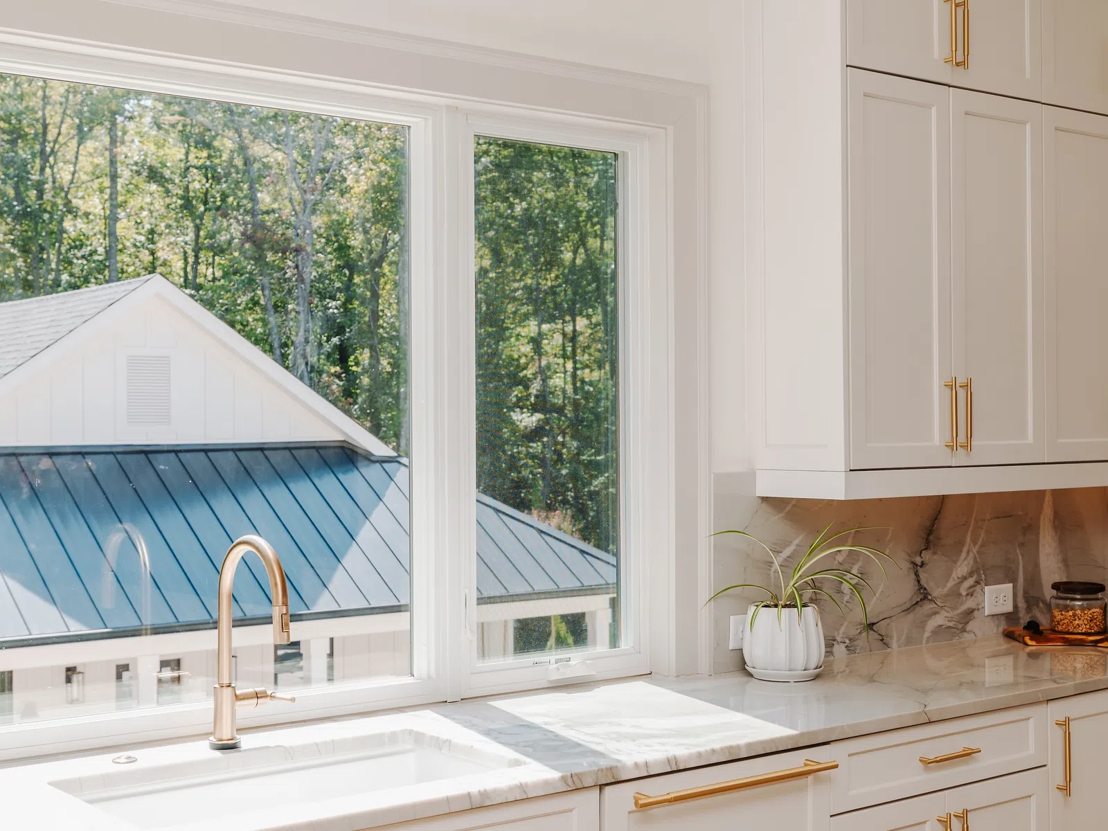 Bright modern kitchen with white cabinets, marble countertops, gold hardware, and large window overlooking trees.