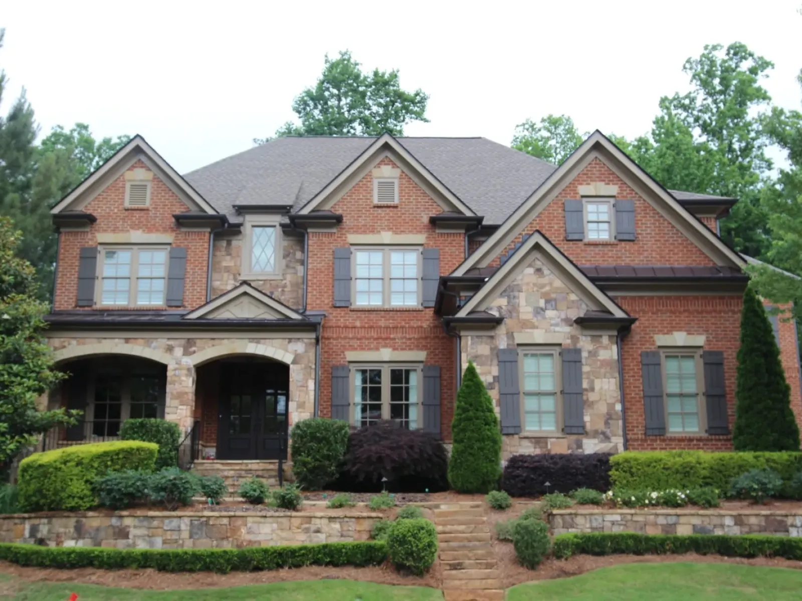 Large two-story house with brick and stone exterior, multiple windows, front porch, and landscaped yard with bushes and trees