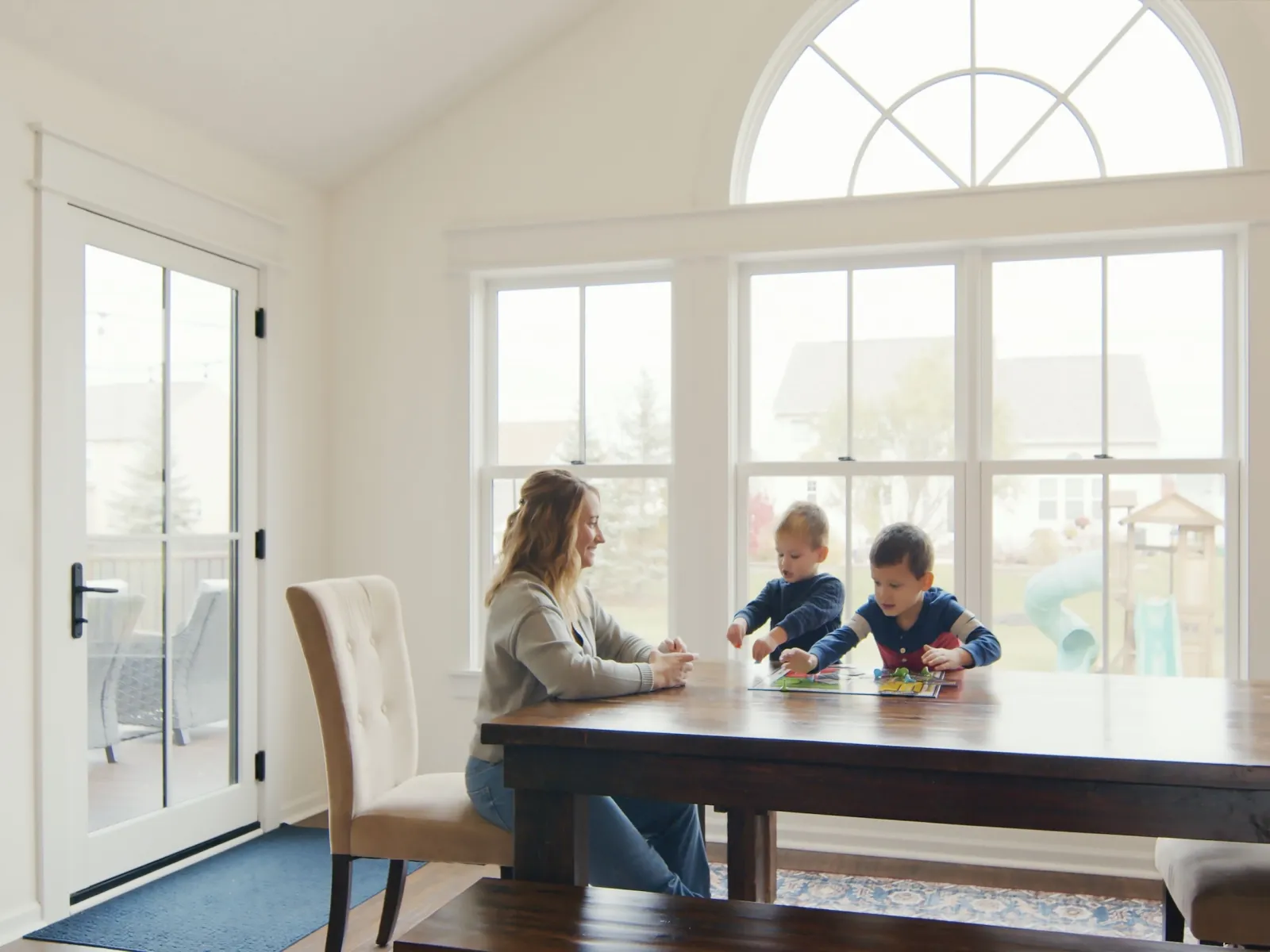 Mother and two young children playing board games at a wooden table in a bright modern dining room.