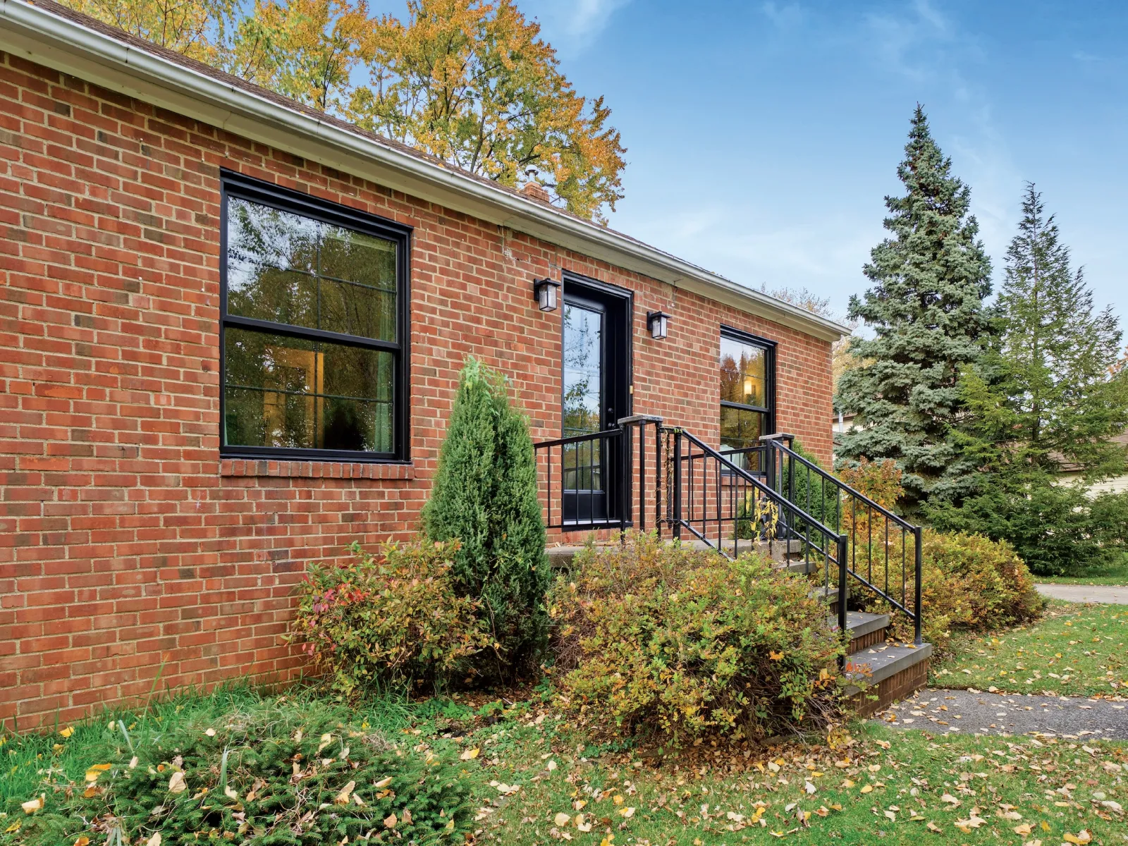 Brick house exterior with black-framed windows, a black door, stairs, and autumn foliage in the yard