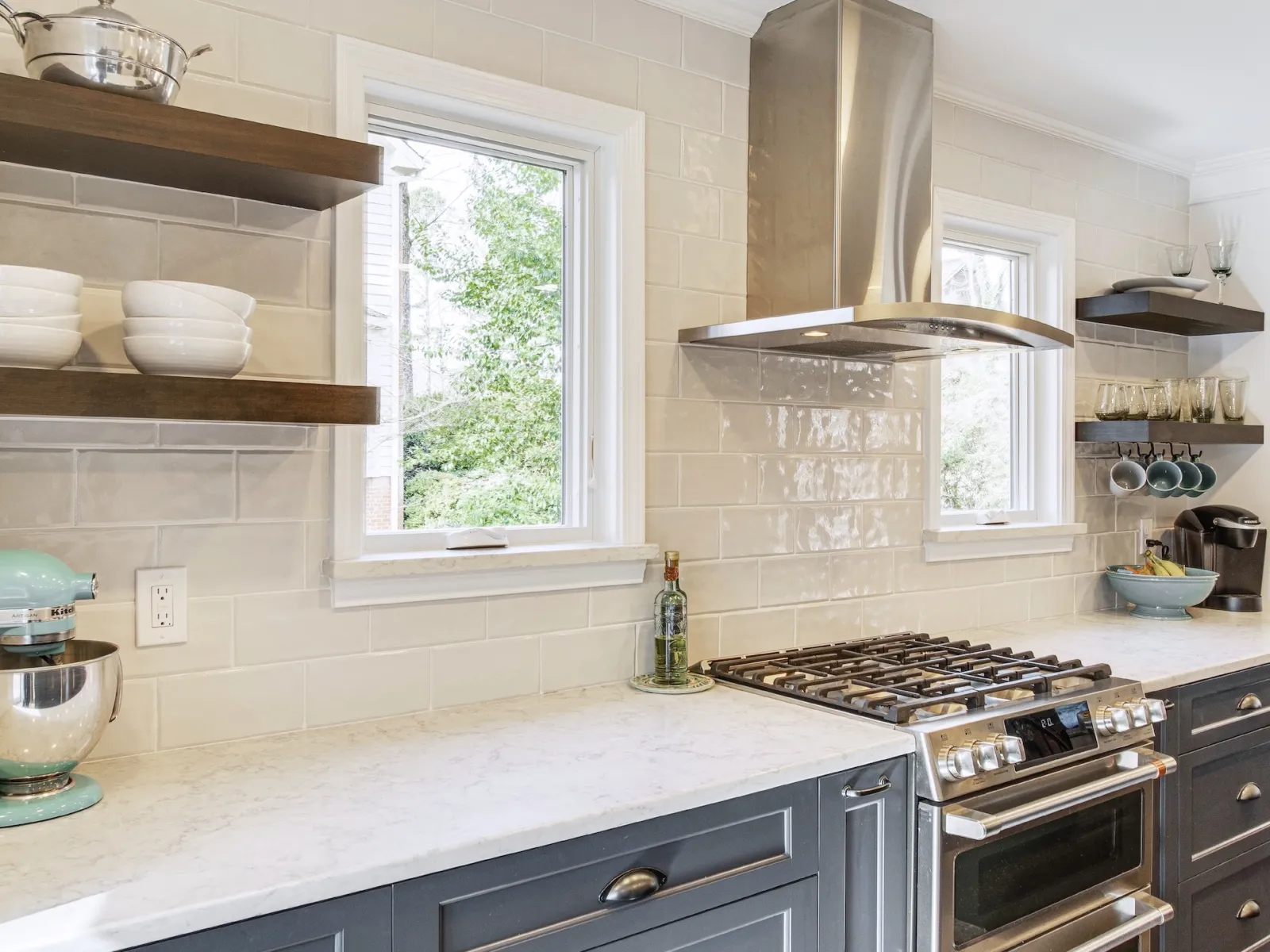 A kitchen with white subway tile on the walls, blue lower cabinets with a marble counter, stainless steel range and vent hood with a white vinyl casement window on either side for ventilation