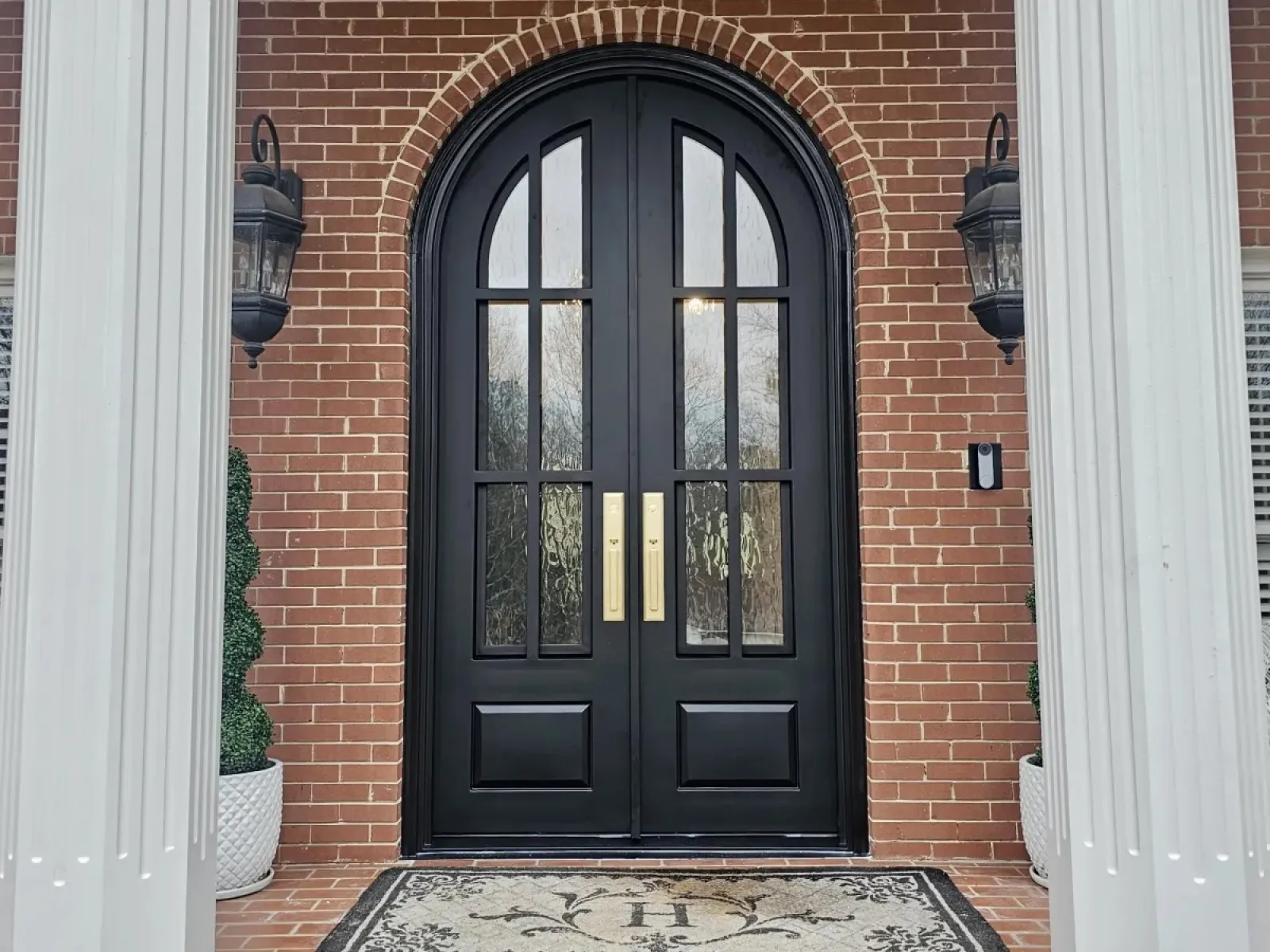 Elegant black double front doors with arched glass panels, brick exterior, white columns, and decorative doormat.