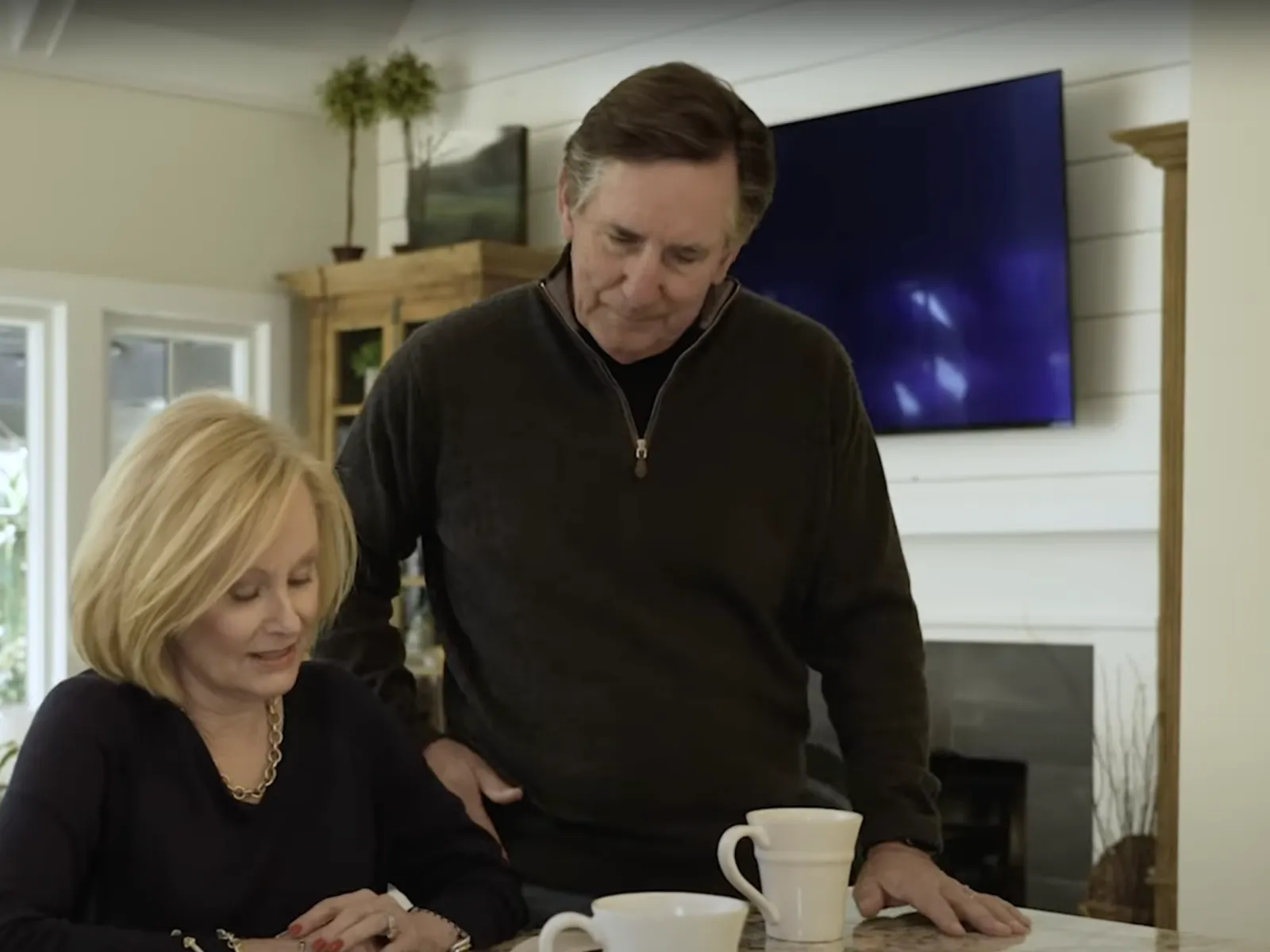 Mature couple in a cozy kitchen, woman sitting and writing while man stands beside her with warm interaction.