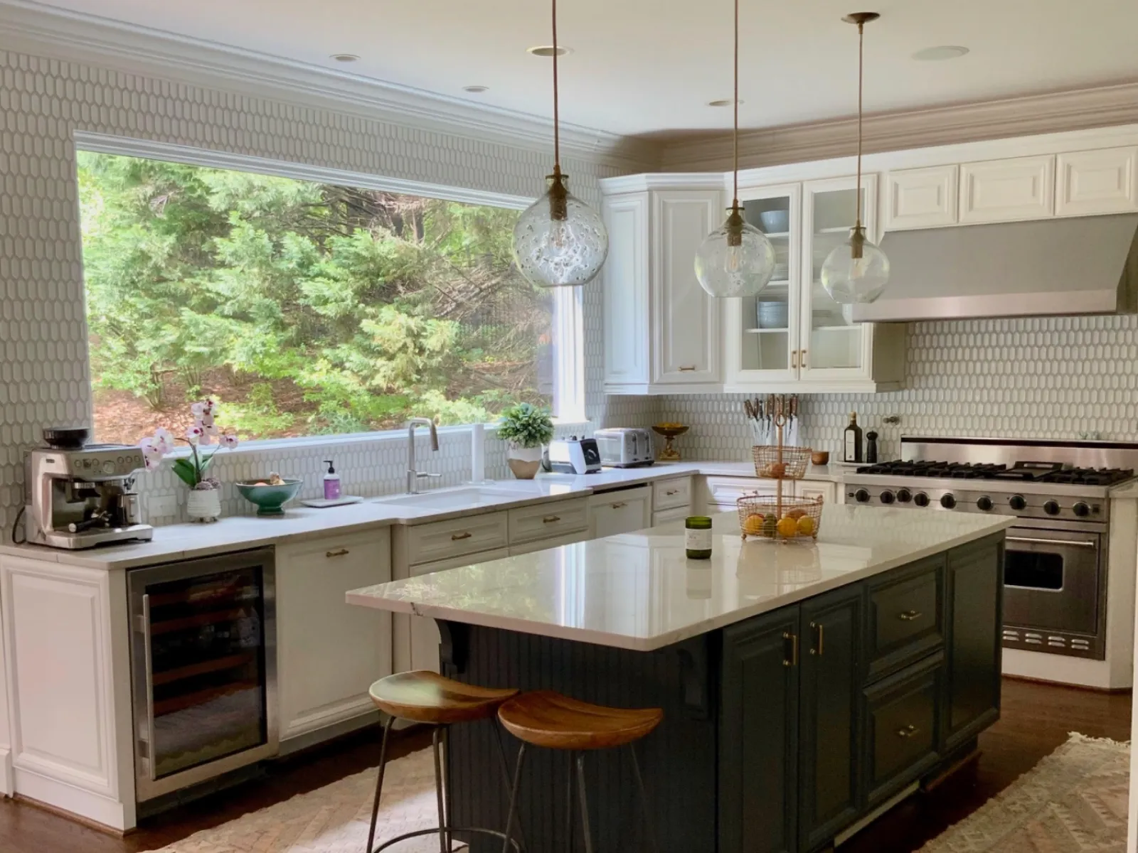 Modern kitchen with white cabinets, dark island, pendant lights, and large window overlooking greenery.