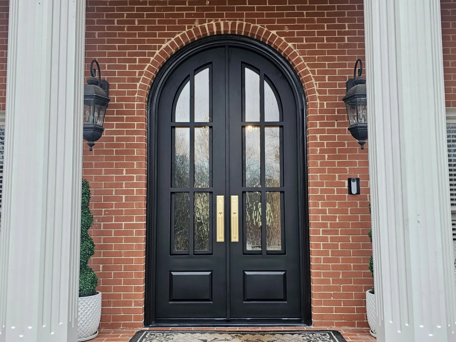 Black arched front exterior doors with brass hardware installed on brick house's columned entry