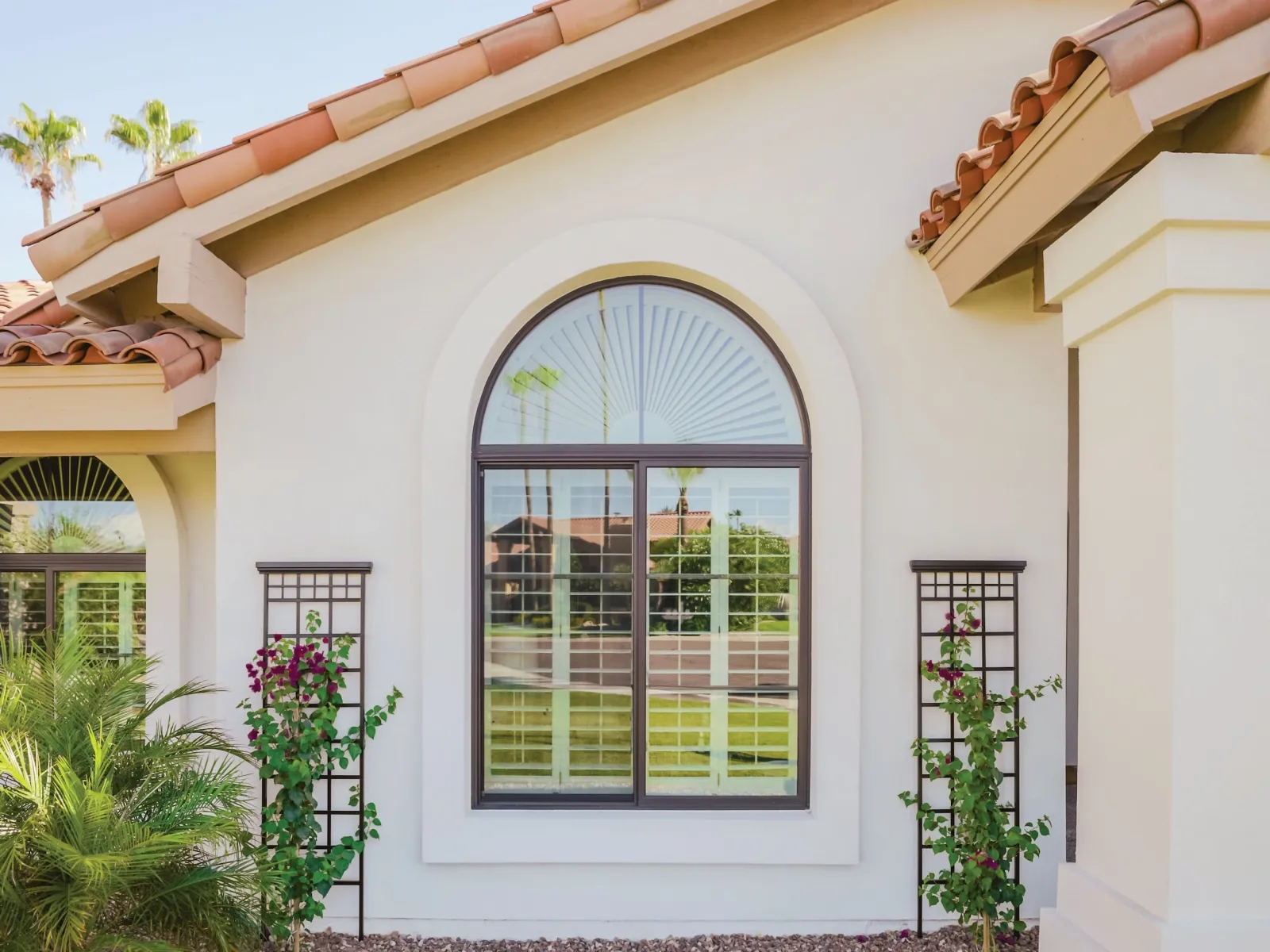 Sunny exterior of a beige stucco house with arched window and terracotta roof tiles surrounded by desert landscaping.
