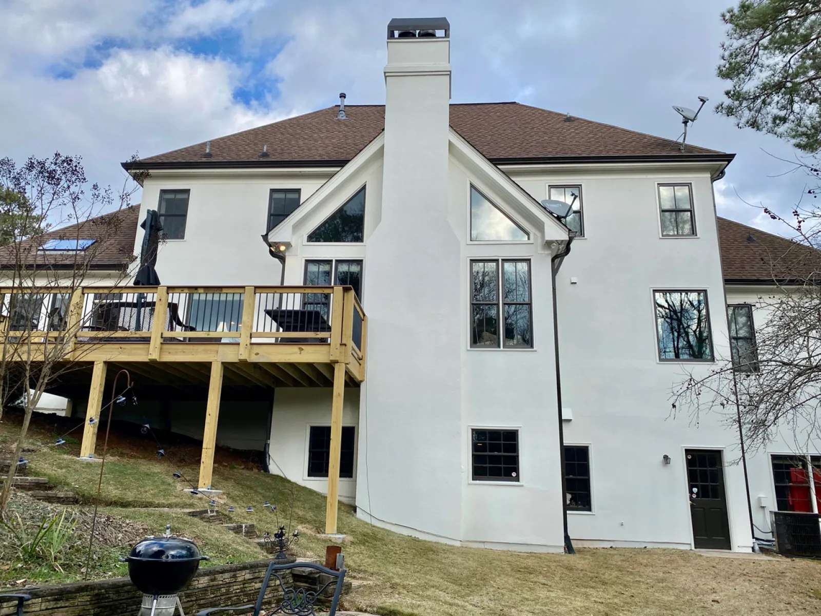 White two-story house with brown roof, large central chimney, wooden deck, and backyard grill on a grassy slope.