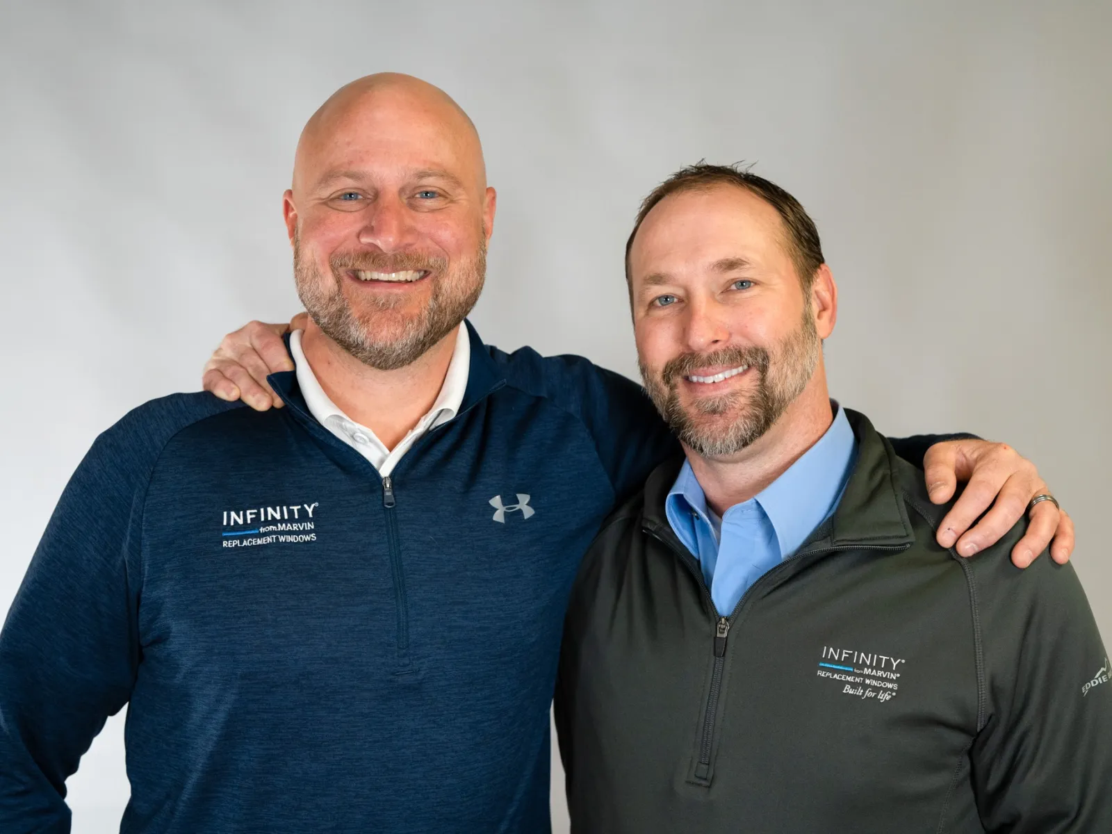 Two smiling men in Infinity branded jackets pose together against a gray background, showing friendly camaraderie.