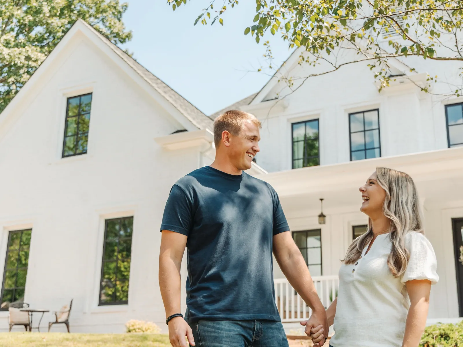 Happy couple holding hands outside their modern white house on a sunny day with greenery around