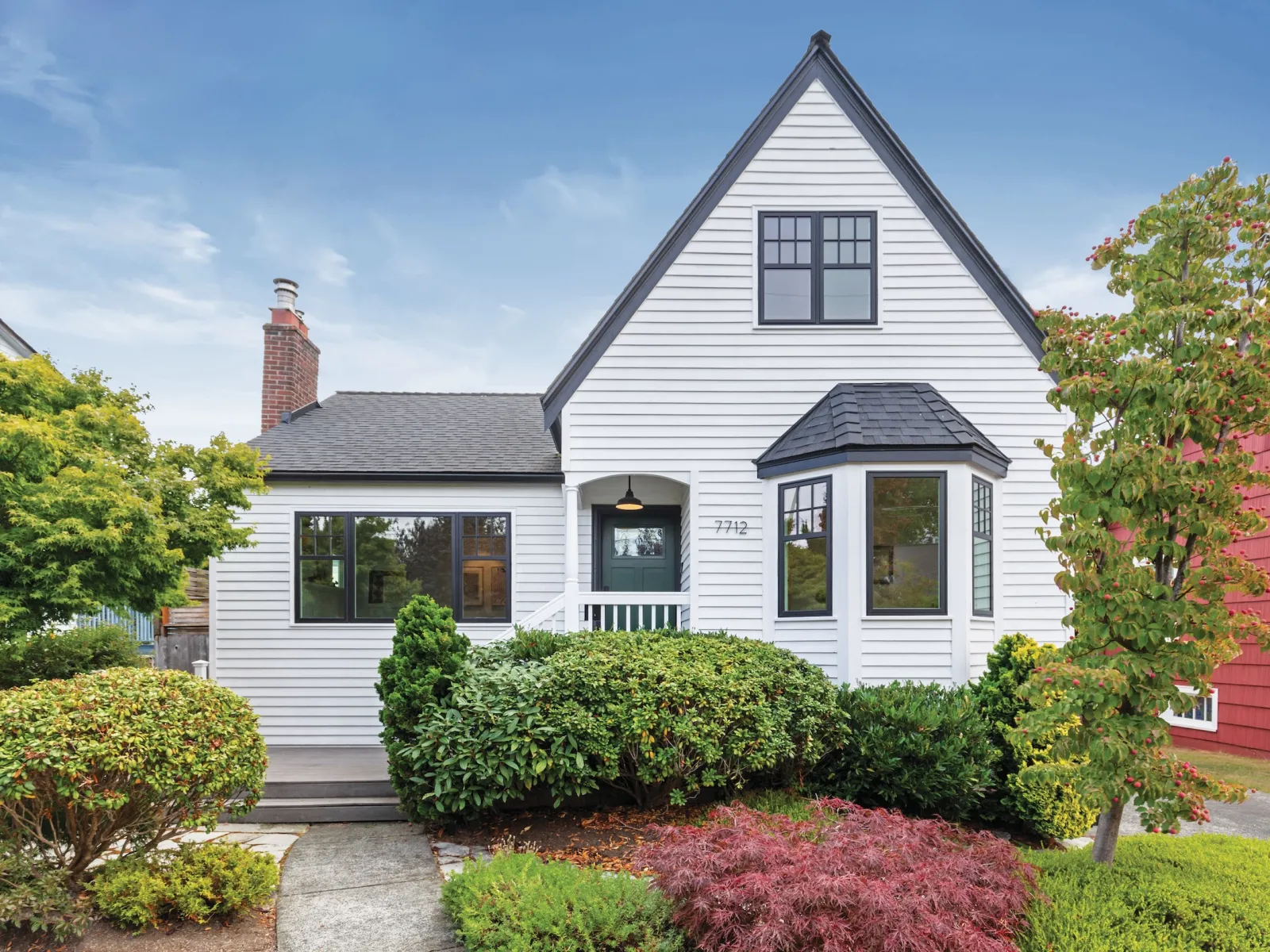 Charming white cottage with steep roof, bay window, chimney, and lush green landscaping under a clear blue sky