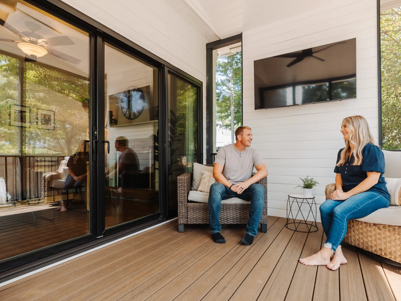 Couple sitting and chatting on a modern outdoor wooden deck with wicker furniture and large glass doors.