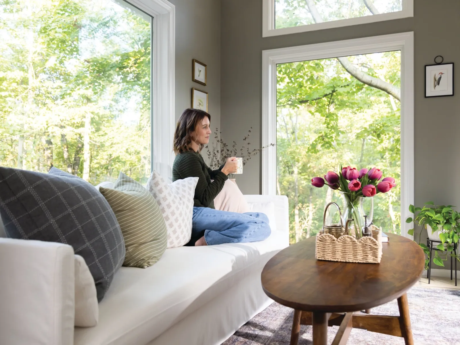 Woman relaxing on white sofa with cushions, holding coffee, beside wooden table with flowers, large windows overlooking trees.