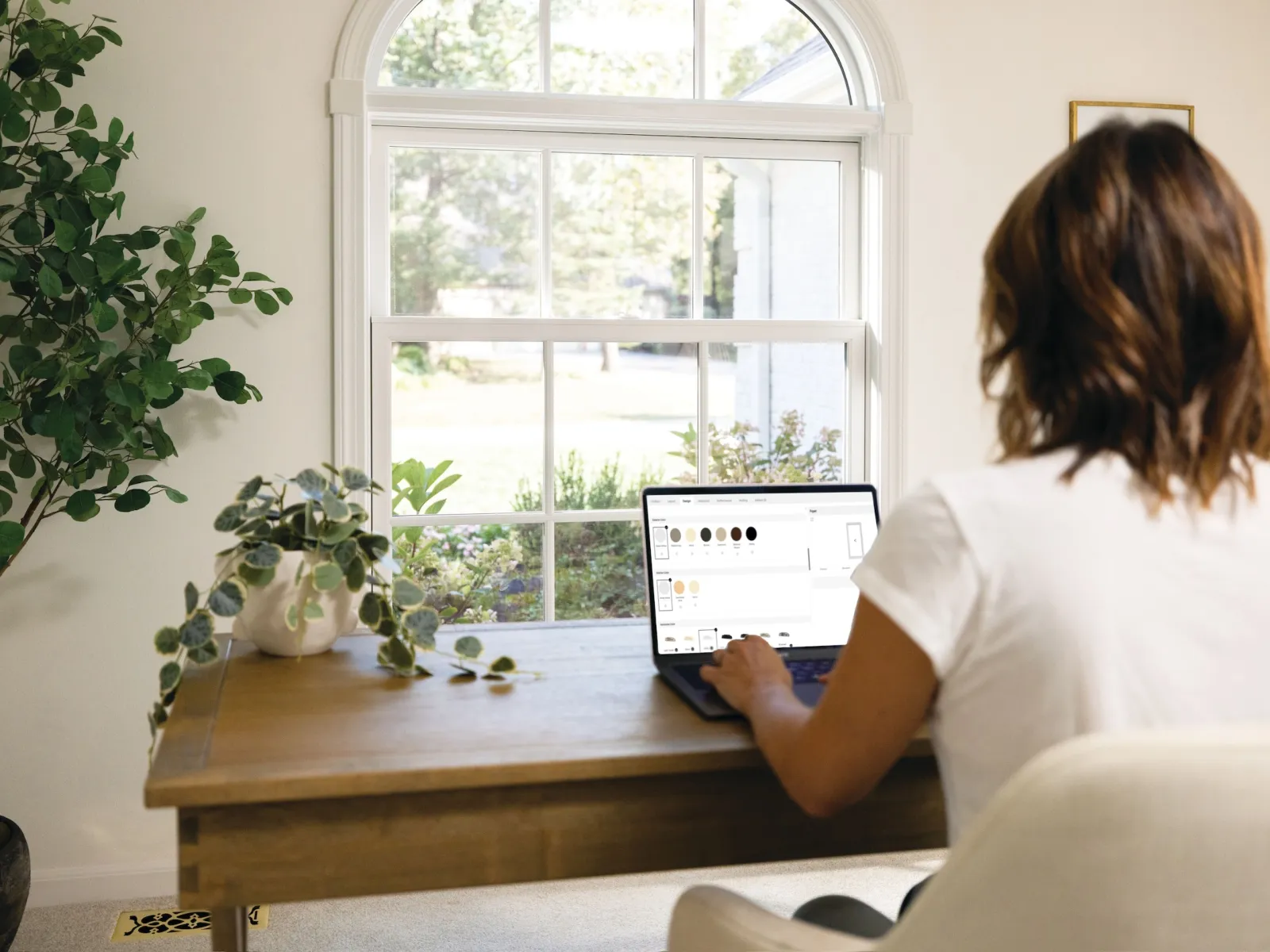 Woman working on laptop at wooden desk near window with greenery and plants in bright home office.