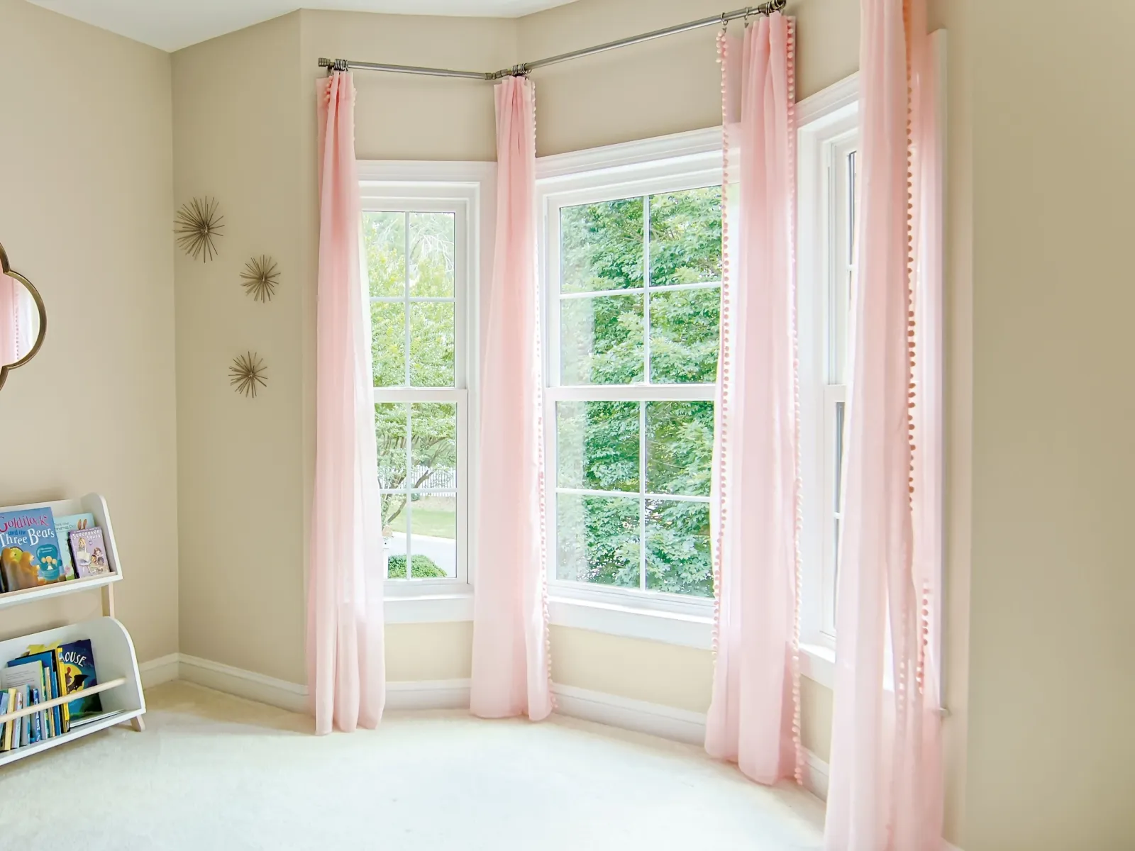 Bright corner room with large windows, sheer pink curtains, children's bookshelf, and beige walls with decor.