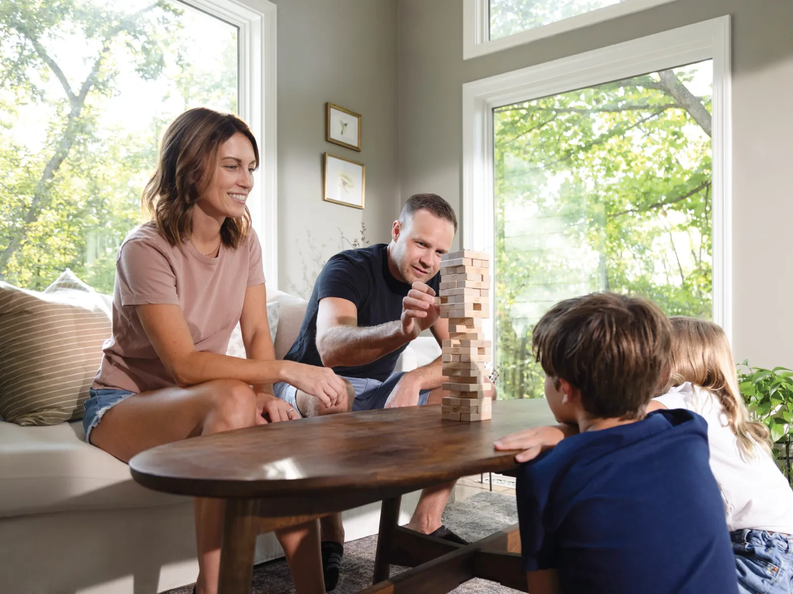 Family of four playing Jenga together on a wooden table in a bright living room with large windows.
