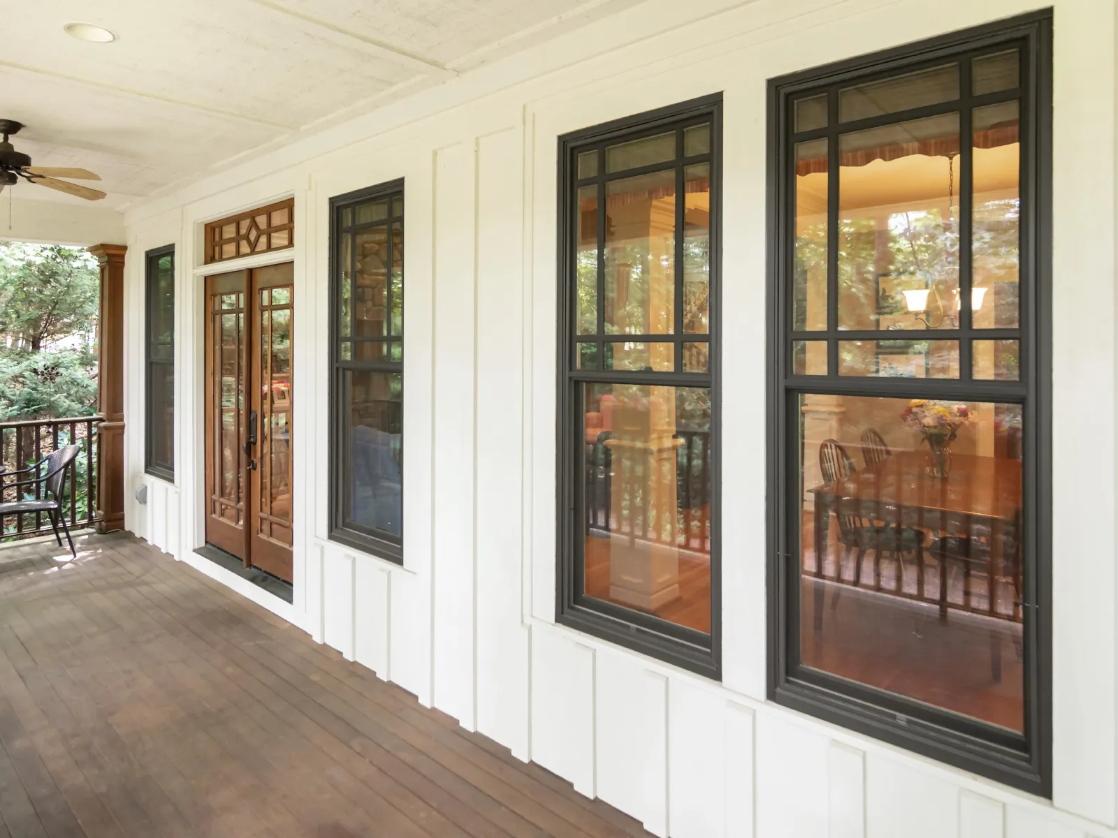 Covered porch with wooden floor, ceiling fan, black framed windows, and dining room visible inside.