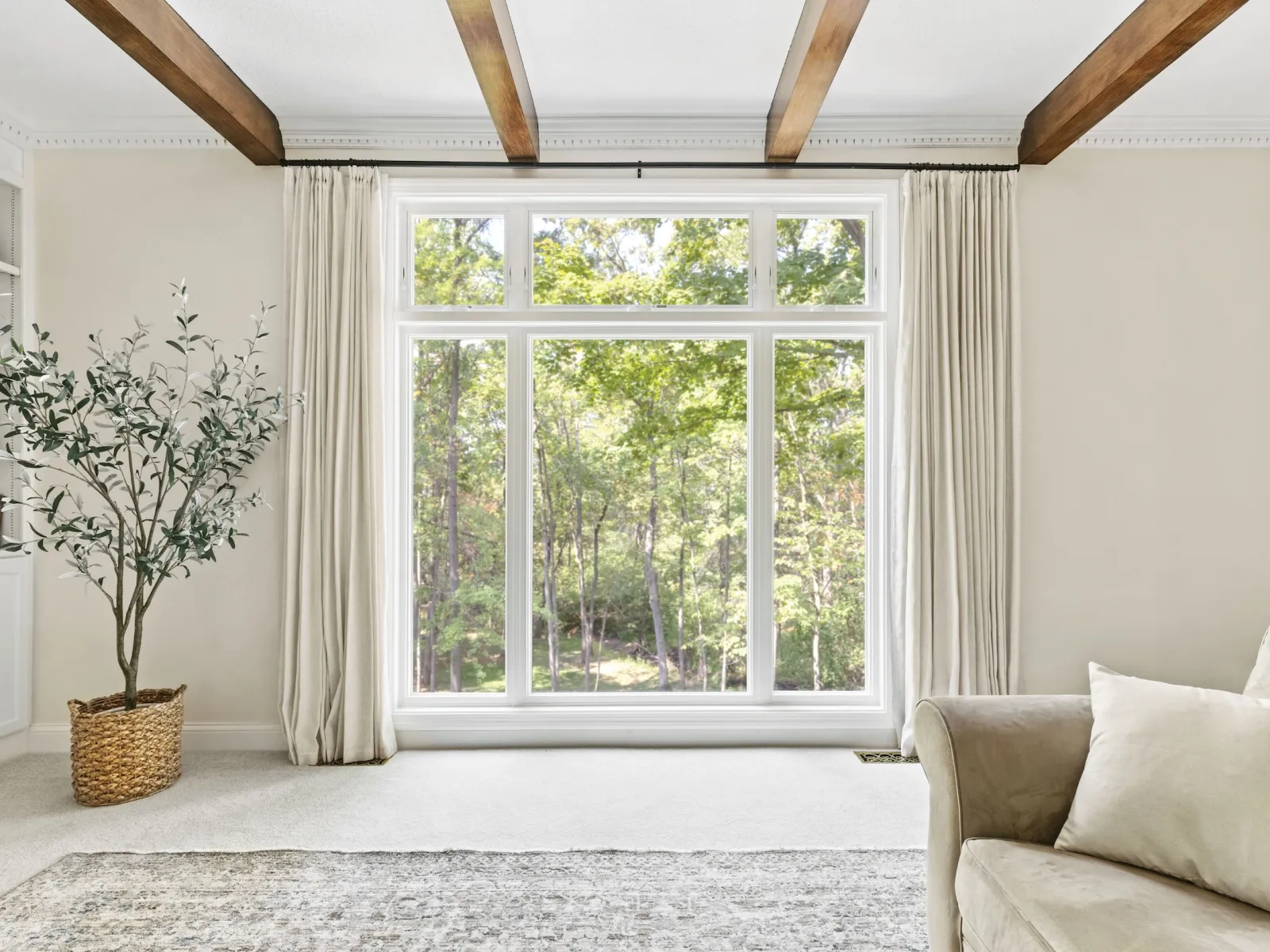 Bright living room with large window, beige sofa, wooden ceiling beams, and a potted plant by the window.