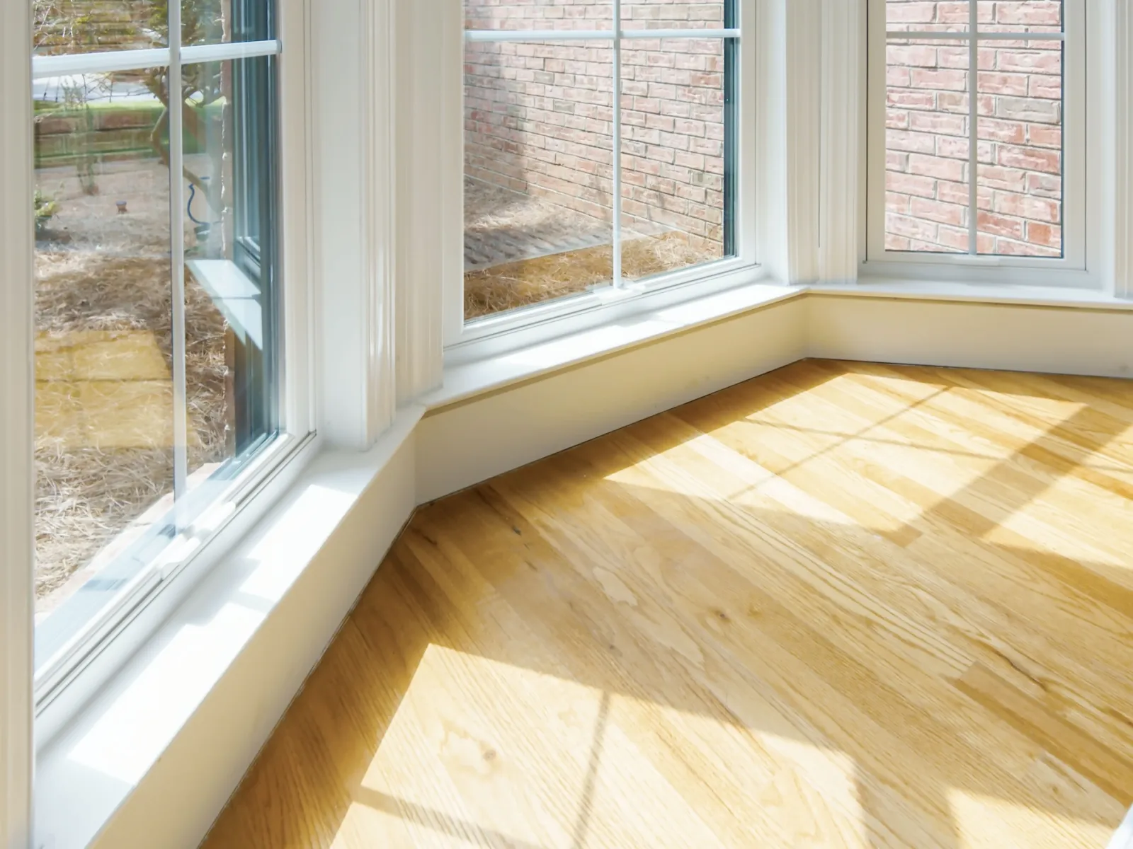 Sunlit hardwood floor by large white-framed bay windows with outdoor brick wall view in a bright room.