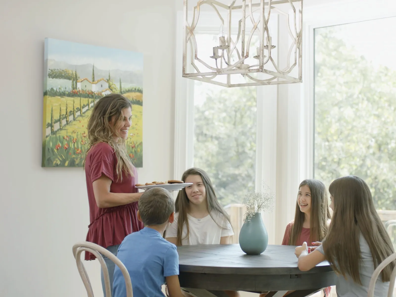 Woman serving cookies to four children sitting around a dining table in a bright room with large windows.
