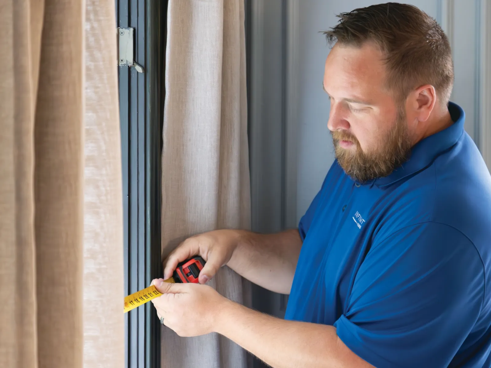 Man in blue shirt measuring window frame using a yellow tape measure by beige curtains.