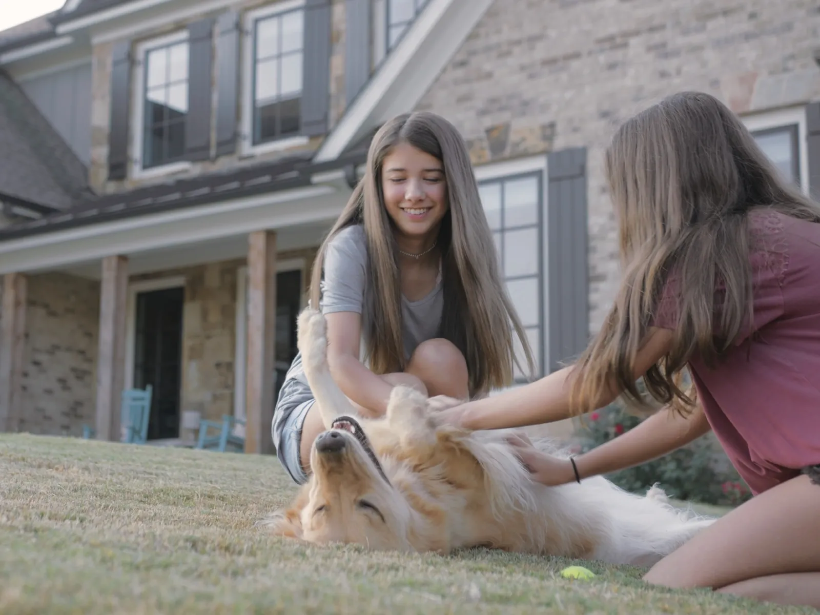 Two girls playing and petting a golden retriever lying on grass in front of a suburban house.