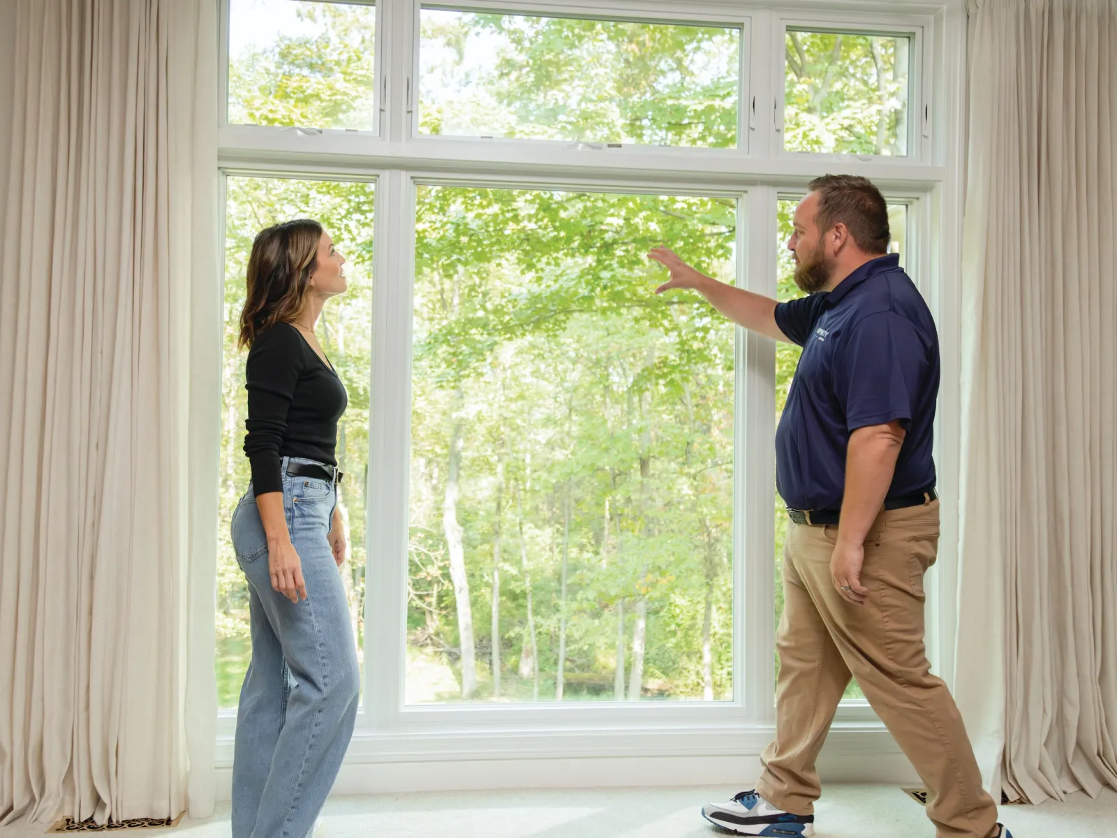 Man explaining window features to woman standing by large clear window with green trees outside.