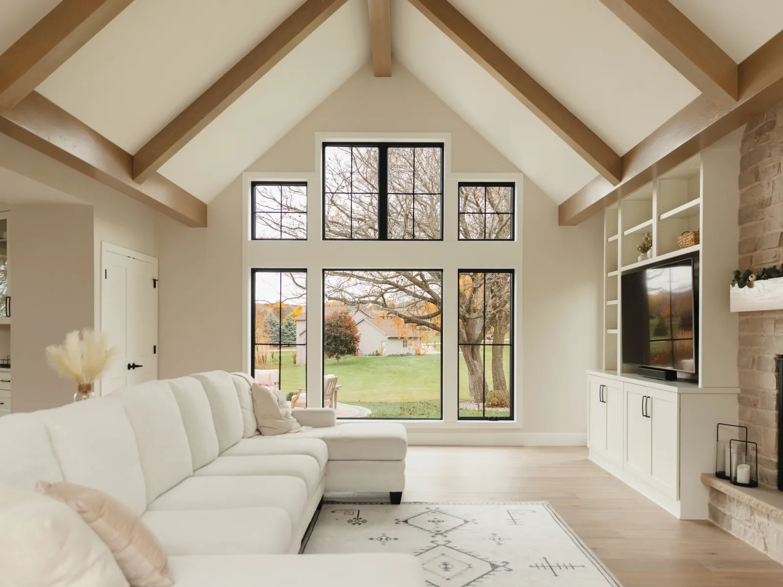 Bright living room with large black-framed windows, white sectional sofa, wood beams, stone fireplace, and built-in shelves.