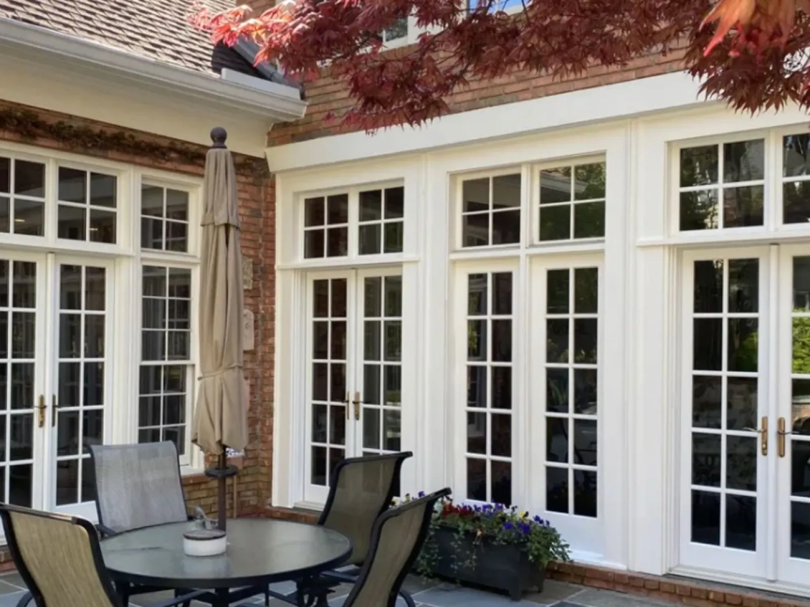 Outdoor patio with round table, chairs, umbrella, large white French doors, and red leafy tree.