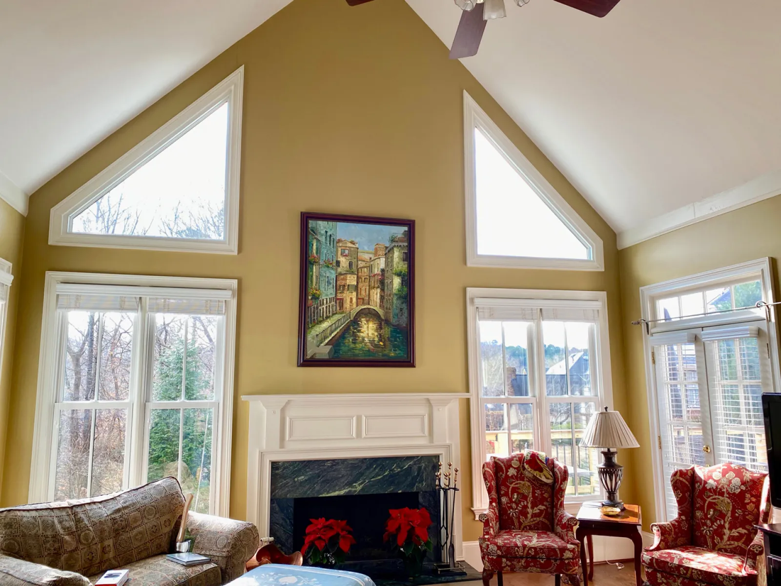 Bright living room with vaulted ceiling, large windows, fireplace with poinsettias, and patterned armchairs.
