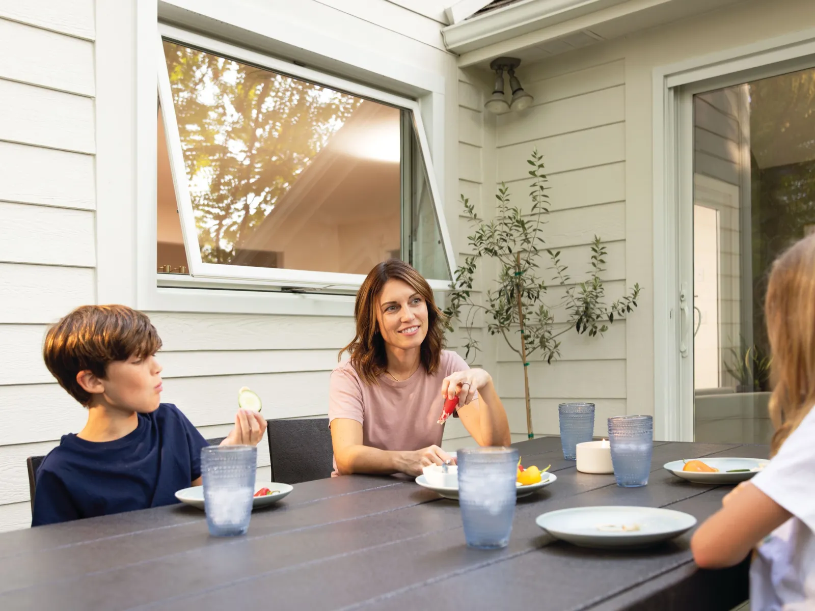 Family of three enjoying a meal and conversation outdoors on a wooden patio table with sunlight and plants.