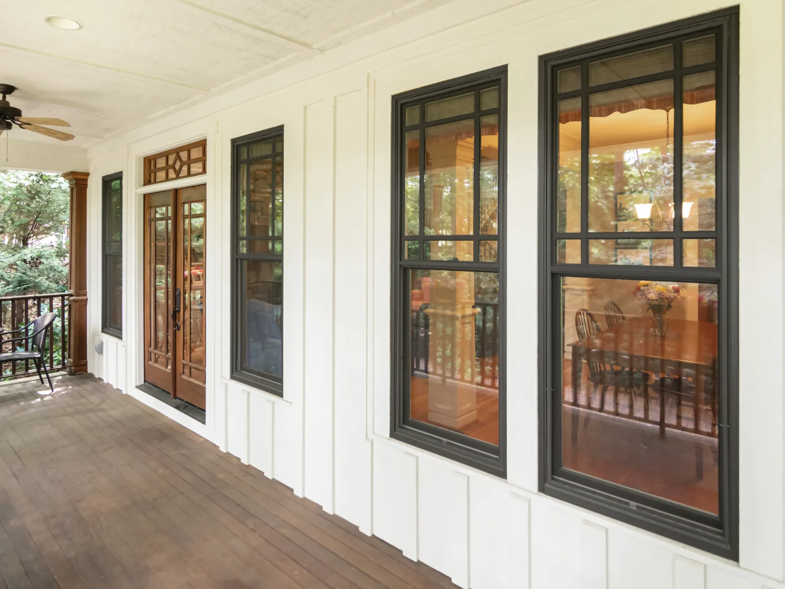 Spacious porch with wooden floor, white walls, dark-framed windows, and wooden double doors leading inside.
