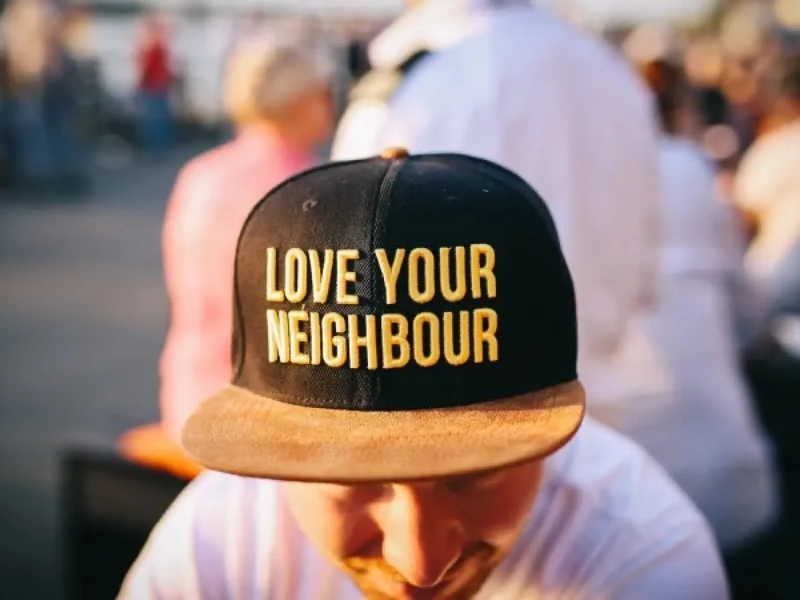 Man wearing a black and brown cap with bold yellow text Love Your Neighbour in an outdoor setting.