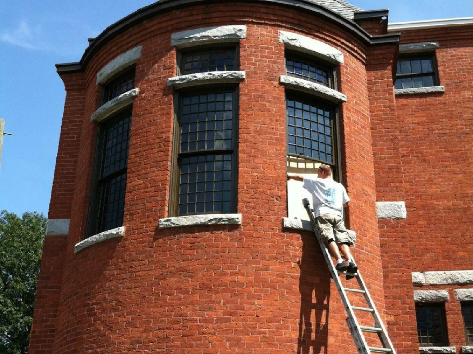 Man on ladder cleaning or painting large curved red brick building windows on a sunny day